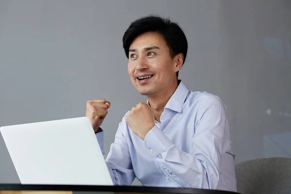 A celebrar o sucesso. Jovem empresário asiático excitado se arma enquanto trabalha no escritório com laptop . — Fotografia de Stock