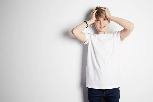 Niño sonriente con camiseta blanca posando frente a una pared blanca y vacía. Niño sonriente posando, pared en blanco en el fondo. Concepto de estilo y moda infantil — Foto de Stock