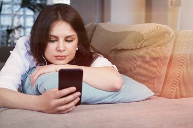 Mutlu Genç esmer kadın oturma odasında kanepede rahatlatıcı akıllı telefon kullanarak