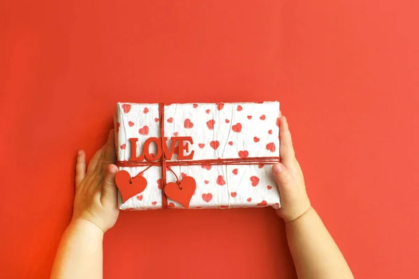 Les mains de l'enfant tenant belle boîte cadeau sur fond rouge. Vacances, vue lumineuse et festive, terrain plat. Bannière de salutation Saint-Valentin. Fête des pères, fête des mères ou fête d'anniversaire . — Photo