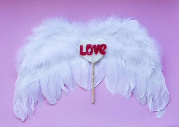 Lollipop op een hart gevormd stick leugens op de vleugels van een engel op een roze achtergrond, een plek voor tekst. — Stockfoto