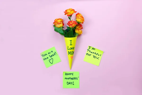 I love mom. Mother's day. Note reminder yellow sticker and beautiful fresh vivid orange roses tinged with red in yellow paper bag on pink background. — Stock Photo, Image