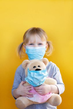 Küçük kız elinde hem tıbbi maske takan bir oyuncak ayı tutuyor. Çocuk karantinada. Koronavirüs ve salgın virüs belirtileri.