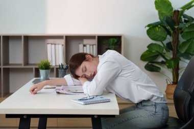 Stresli genç bir kadın, iş hayatı dengesi ve zihinsel sağlığının zorluklarını göstererek başını masaya yaslar..