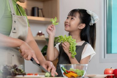 Bir yetişkin renkli bir salata hazırlarken, bir çocuk taze marulun tadına bakarken mutfakta neşeli bir an. Sağlıklı beslenme alışkanlıklarını vurgulamak.