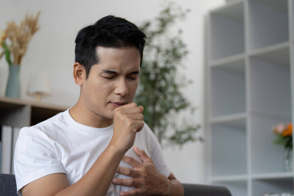 A young man coughs while holding his chest, showing signs of respiratory distress in a modern home setting.