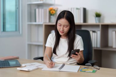 Genç bir kadın akıllı telefonunu kullanarak, etkili finans yönetimi ve kişisel organizasyonunu sergileyerek notlar yazar..