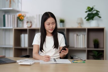 Genç bir kadın defterine akıllı telefonunu kullanarak kişisel finans planlaması ve organizasyonunu vurguluyor..