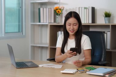 Neşeli genç bir kadın, rahat bir ofiste akıllı telefonunu kontrol ederken gülümsüyor ve dijital bağlantının önemini vurguluyor..