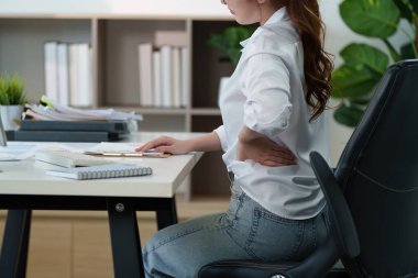Genç bir profesyonel masasında otururken belinde rahatsızlık hissediyor, ergonomik uygulamaların önemini vurguluyor..