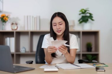 Masasında oturan genç bir kadın, faturalarını ve mali belgelerini incelerken stresli görünüyor. Mali yönetim ve stres kavramı.