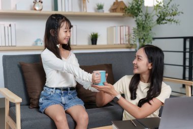 Bir kızın annesine bir kupa uzatması gibi tatlı bir etkileşim ev ortamında bakım ve bağlantıyı sembolize eder..