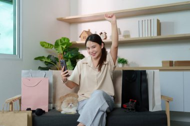 Çevresinde alışveriş torbaları ve rahat bir atmosferle çevrili kedisiyle çevrimiçi alışveriş yapan bir kadın neşe ifade ediyor..