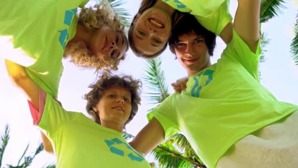 Quatro jovens voluntários em camisetas verdes com uma foto de reciclagem formando dobradiças sob palmeiras na costa de uma praia oceânica, depois de limpar a praia de detritos de plástico. Voluntariado e — Vídeo de Stock