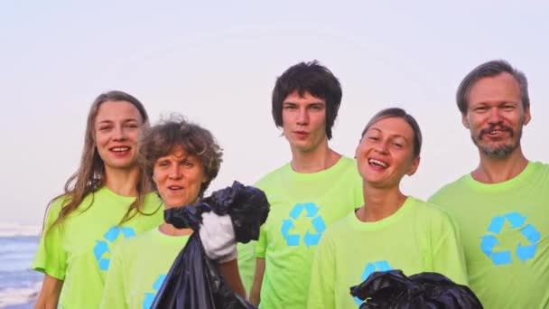 Cinq jeunes volontaires en t-shirts verts recyclés d'images, ramassent les ordures sur une plage océanique, puis posent sur la caméra avec des sacs d'ordures collectées. Concept de bénévolat et recyclage — Video