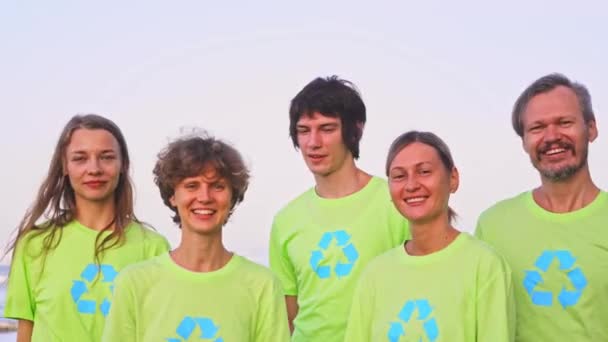 Five young volunteers in green t-shirts with image recycle, collect garbage on an oceanic beach, then pose on the camera with bags of collected garbage. Volunteering and recycling concept — Stock Video