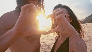 Öpüşmek, el ele tutuşarak ve ağır çekimde bir kalp şekil verme sahilde günbatımı zevk çekici güzel genç seyahat çift. Deniz yaz tatil mutlu romantik turist kavramı.