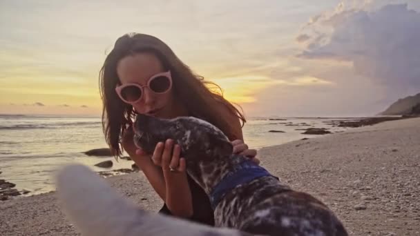 Молодая беззаботная женщина в солнечных очках играет с собакой, гладит и целует собаку на пляже во время удивительного заката. Морские летние каникулы — стоковое видео