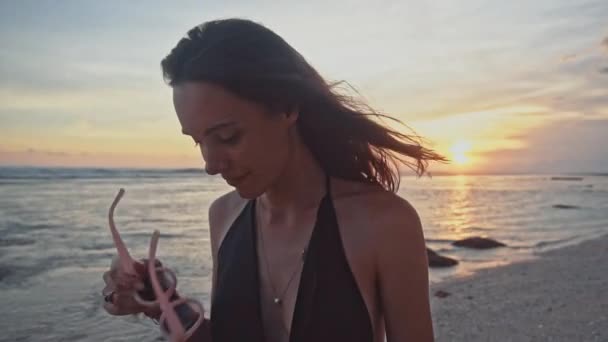 Young attractive woman in black dress and sunglasses looks and smiles at the camera on an empty oceanic beach at sunset. Sea summer vacation happy romantic travel concept — Stock Video