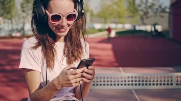 Jeune femme ou adolescente souriante et riante avec smartphone et écouteurs écoutant de la musique dans le parc ou sur le terrain de sport. Concept de technologie, mode de vie et personnes — Video