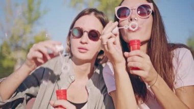Kabarcıklar üfleme ve having fun üzerinde açık havada güneş gözlüğü iki genç kadın portresi. Sinematik ağır çekim, kabarcıklar, yaz yaşam tarzı üfleme Genç Festivali kadın