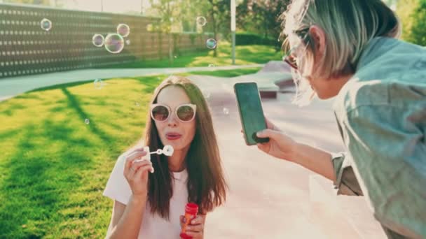 Giovane donna in occhiali da sole soffia bolle e divertirsi e la sua amica sparare al telefono. Cinema al rallentatore, giovane festival donna soffia bolle, stile di vita estivo — Video Stock