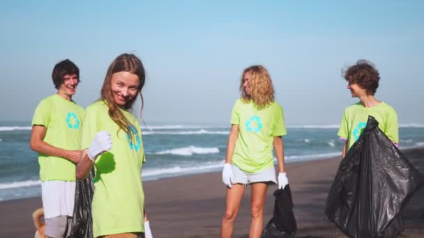 Cuatro voluntarios en camisetas verdes con reciclaje de imágenes recogen basura en la playa, mirando a la cámara con bolsas de basura recolectada. Concepto de voluntariado y reciclaje. Espacio de copia de conciencia ambiental — Vídeo de stock