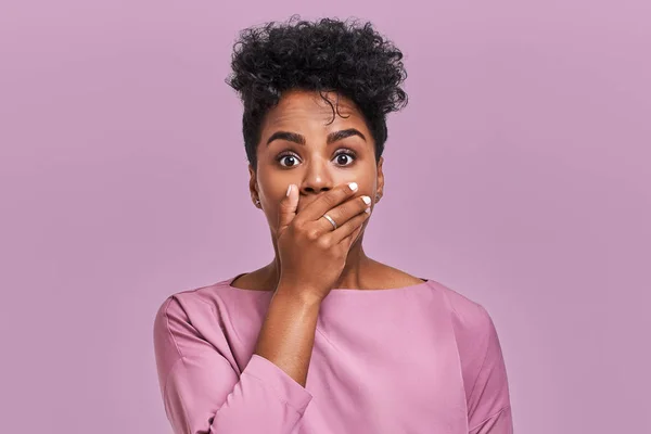 Horizontal shot of surprised African American woman covers mouth with both hands, tries to be speechless, looks with shocked expression, notices something strange, isolated over lavender blank wall