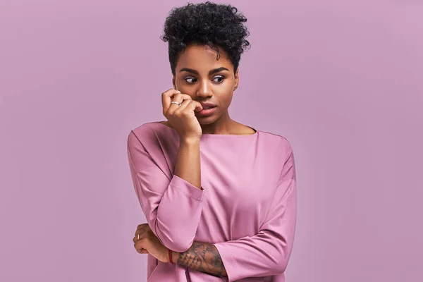 Embarrassed African American woman pursed lips, looks with puzzlement, hears something not clear, dressed in casual top, isolated over lavender background. People and facial expressions