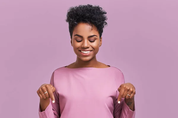 Engraçado surpreendido afro-americano feminino olha com olhos alegres para baixo e indica como mostra algo, vê coisas cômicas, isolado sobre parede de lavanda. Positivo espantado jovem posa no estúdio — Fotografia de Stock