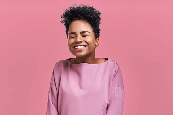 Retrato de felicidade alegres afro-americano feminino, amplamente sorriu e fecha os olhos, celebra seu sucesso, poses contra fundo rosa. Pessoas, felicidade, conceito de sucesso — Fotografia de Stock