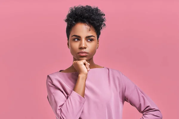 Muhteşem koyu tenli Genç kadın, Afro saç modeli ve kendine güvenen bak, teşkil etmektedir moda dergisi, görünüyor bir kenara düşünceli bir poz veriyor pembe studio arka planı