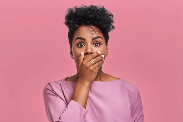 Horizontal shot of surprised African American woman covers mouth with both hands, tries to be speechless, looks with shocked expression, notices something strange, isolated over pink blank wall