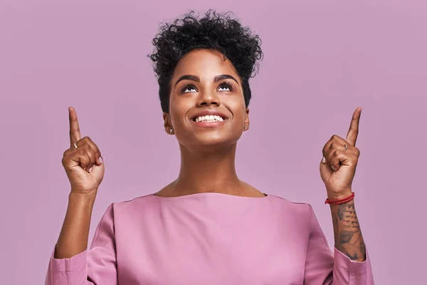 快乐漂亮的非洲裔美国女性用食指向上表示 有友好的微笑 深色的皮肤 卷曲的头发 在薰衣草背景下显示出空白 广告概念 — 图库照片
