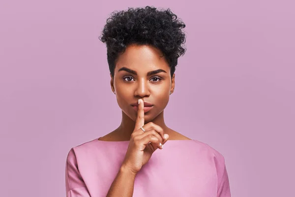 Terrific dark skinned girl keeps fore finger on lips, has mysterious expression, shows silence gesture, wears casual striped top, headband, isolated over lavender background. Dont make noise, please — Stock Photo, Image