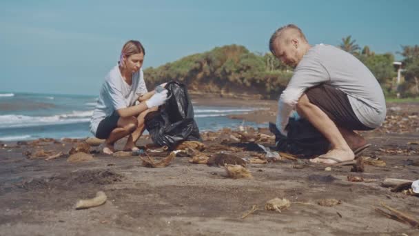 Deux bénévoles nettoient sur la plage. Nettoyants ramassant les ordures sur la plage de sable noir dans un sac en plastique noir. Ranger les ordures sur la plage. Concept de sensibilisation environnementale — Video