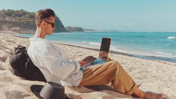 Młody miejski cyfrowy Nomada tysiąclecia siedzi na plaży i działa na laptopie — Wideo stockowe