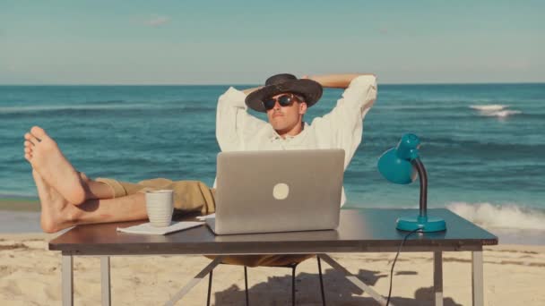 若い男がノートパソコンを持ってビーチに座り、テーブルの上に足を置いて休んでいる — ストック動画