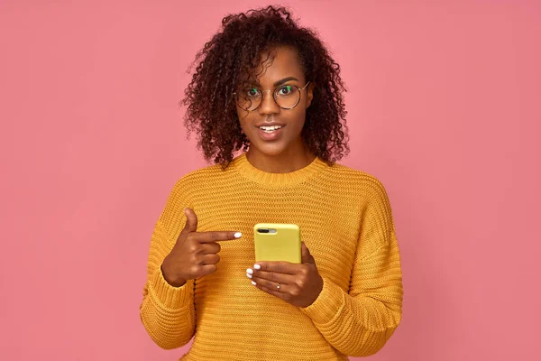 Joyous African American Lady met grote glimlach, punten op het scherm van de mobiele telefoon, verheugt zich over nieuwe app, geniet van online communicatie, in hoge geest, geïsoleerd op roze achtergrond. Technologie concept — Stockfoto