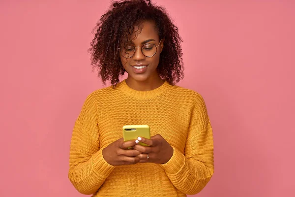 Tevreden mooie zwarte vrouw houdt moderne mobiele telefoon, teksten met vriend online in de chat, in een oranje trui, poses tegen roze muur, verbonden met High Speed Internet. Shot van tiener met apparaat — Stockfoto