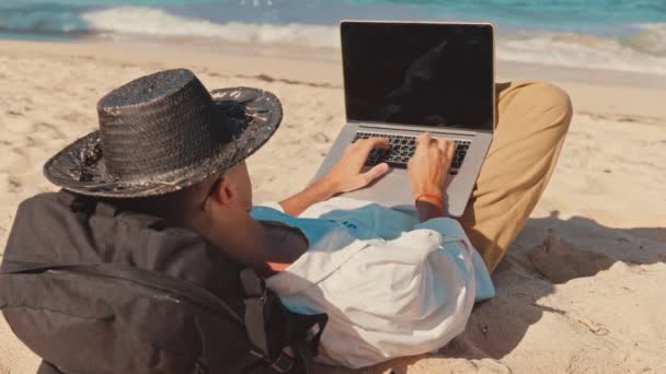 Primer plano detrás del hombro de un joven profesional freelancer o turista, tumbado en la playa descalzo, trabajar de forma remota desde la oficina durante las vacaciones. Concepto carrera remota, habilidad de profesión digital en línea — Vídeos de Stock