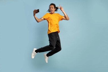 Tam uzunlukta yan profil vücut boyutu fotoğraf funky komik genç Afrikalı-Amerikalı adam yüksek atlama, v-işareti jest göstermek, selfie çekmek yapmak, turuncu kıyafet giyer, güzel bir gün tadını çıkarın. Yalıtılmış mavi arka plan.