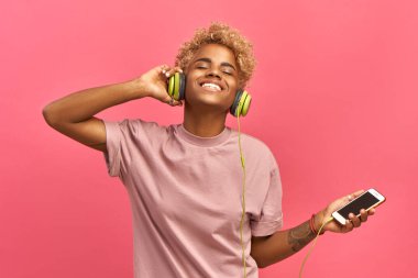 İnsanlar, müzik, duygu konsepti. Afro saç stili, melodiyle dans eden, gözleri kapayan, kulaklıkla yüksek sesle şarkı dinleyen, akıllı telefon kullanan zevkli bir kadın. Pembe renk galip geliyor..