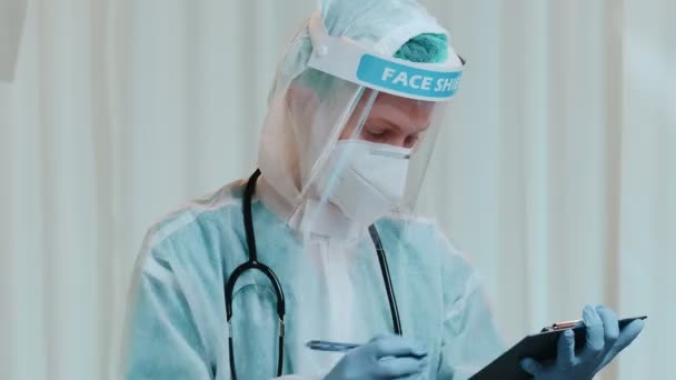 Vrouwelijke arts met Hazmat, beschermend masker en gezichtsschild schrijven patiëntennotities — Stockvideo