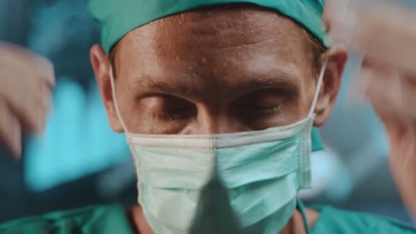 Moe ervaren mannelijke arts met beschermend masker op zijn gezicht en chirurgische pet — Stockvideo