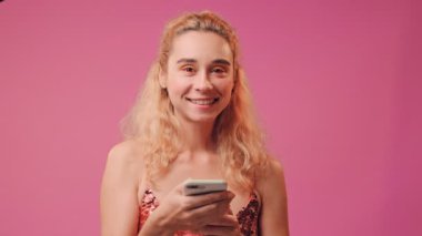 Mutlu kız cep telefonunu tutuyor ve pembe arka planda iyi bir jest yapıyor..