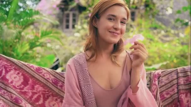 Ung kvinna ler, poserar med rosa blomma på hängmatta i tropisk trädgård — Stockvideo