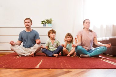 Anne, baba ve iki anaokulu öğrencisi birlikte yoga yapıyorlar. Aileler hafta sonu evde karantinada negatif duyguları rahatlatmak için meditasyon yaparlar..