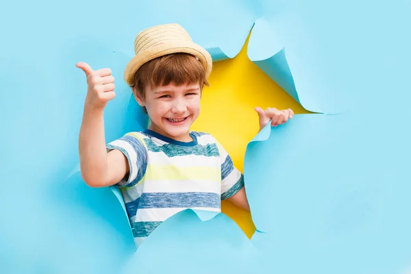 Lindo niño pequeño en un sombrero muestran los pulgares hacia arriba y posa a través de un agujero de papel roto . —  Fotos de Stock