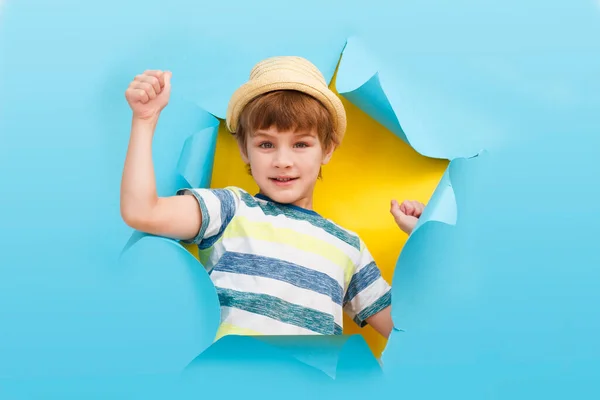 Netter kleiner Junge mit Hut zeigt Daumen hoch und posiert durch zerrissenes Papierloch. — Stockfoto